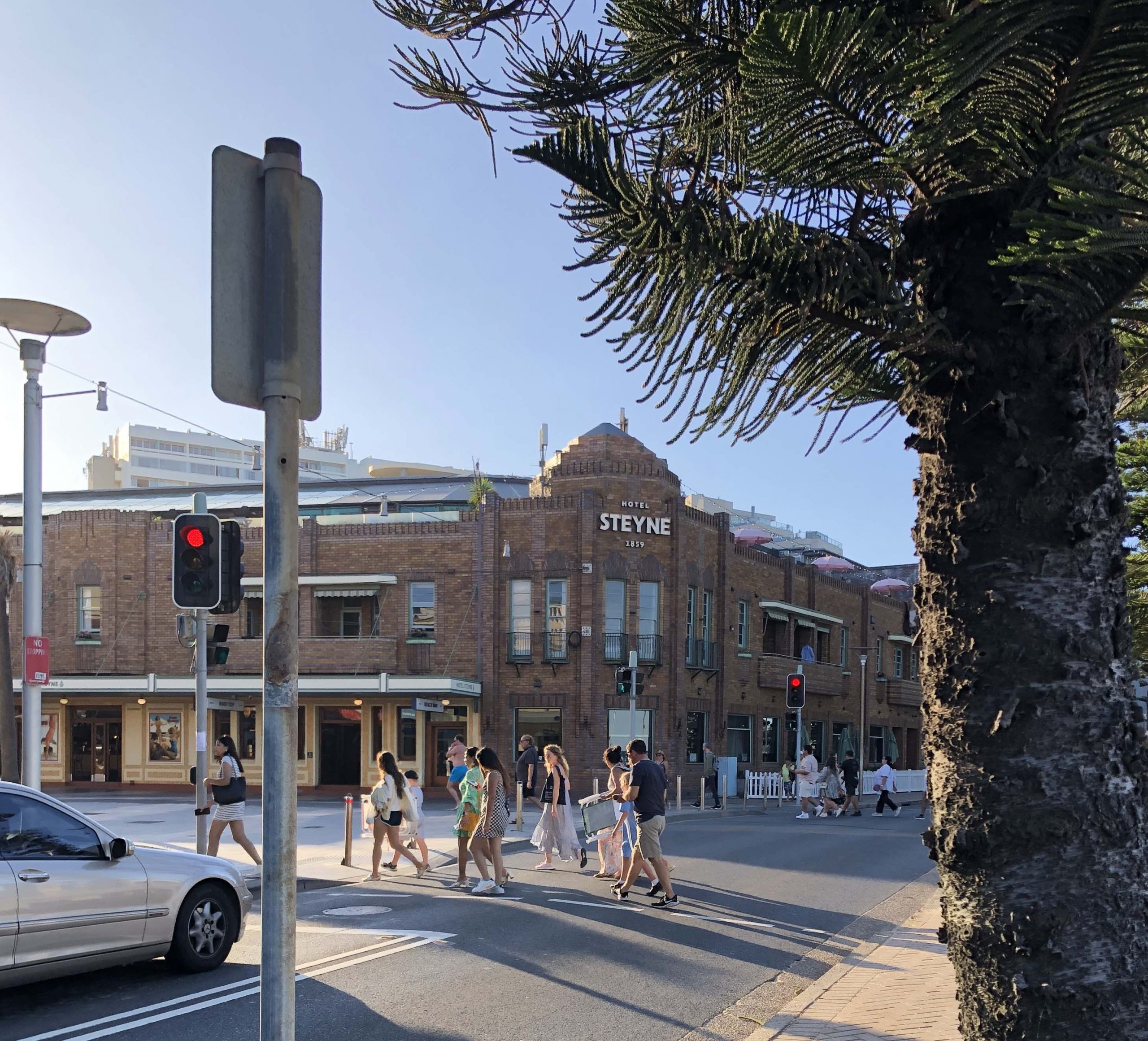 Street view of the the Hotel Steyne , built in 1859. People crossing at traffic lights and the hotel. A two level brick building overlooking The Corso - street running along sidethe  Manly beach .